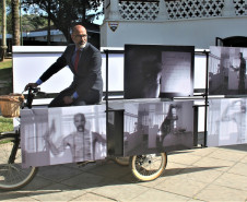 Obra "A Dúvida da Verdade" de Sérgio Adriano H., composta por uma bicicleta triciclo com caixa baú, estrutura metálica modular e 12 fotografias impressas sobre PVC.