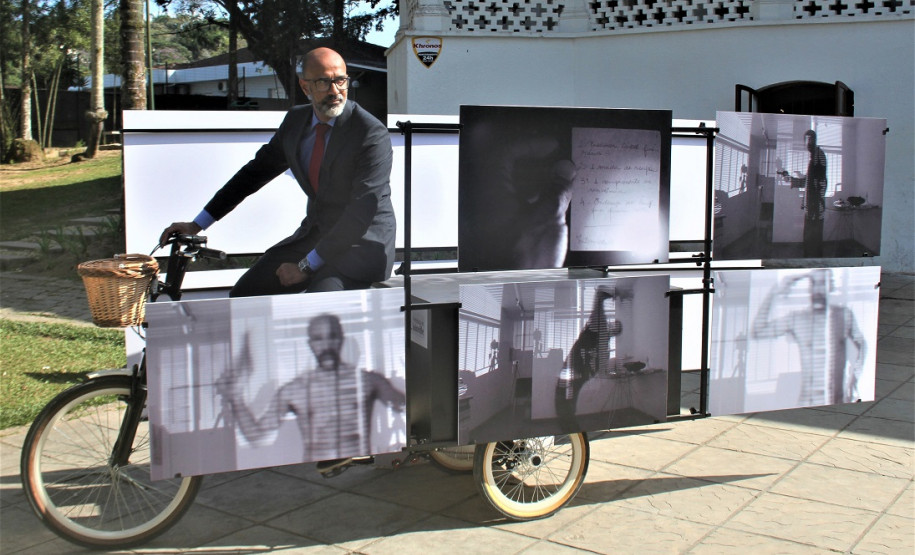 Exposição Arte e Vida Obra "A Dúvida da Verdade" de Sérgio Adriano H., composta por uma bicicleta triciclo com caixa baú, estrutura metálica modular e 12 fotografias impressas sobre PVC.